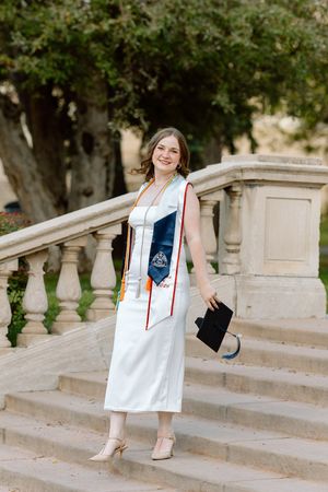 Young woman in white graduation dress holding cap on stone steps, wearing academic stole outdoors.
