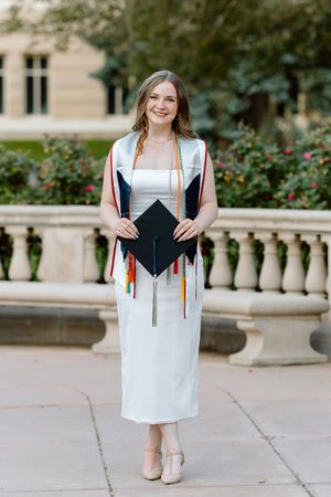 Smiling graduate in white dress holds cap with colorful tassels outdoors at a university campus.