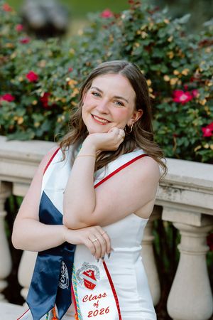 Smiling graduate wearing Class of 2026 sash posing near stone railing with red roses in background.
