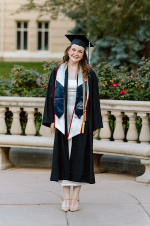 Smiling graduate in black cap and gown with honor stoles poses outdoors on campus walkway.