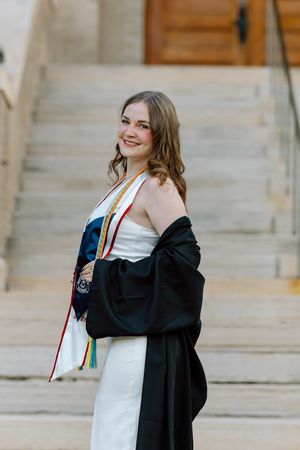Smiling graduate in white dress holds black gown with colorful honor stoles on stone steps.