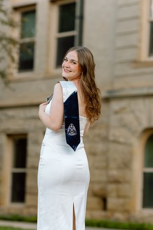 Graduate in elegant white gown with navy blue graduation stole poses outdoors at university campus.