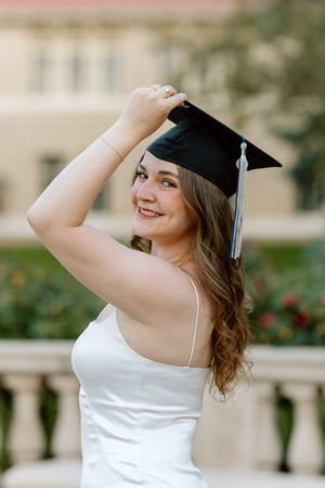 Smiling young woman in white dress holding black graduation cap outdoors near a classic building.