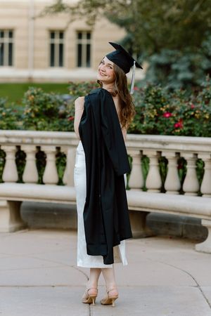 Smiling graduate in black cap and gown over white dress posing outdoors at university campus.