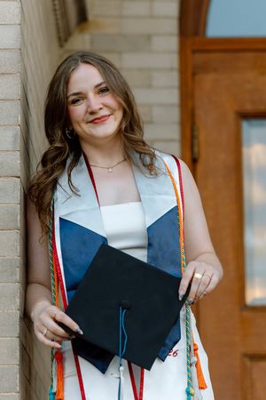 Smiling college graduate holding black cap wearing white dress and colorful graduation stoles outside.