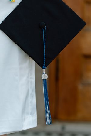 Close-up of a black graduation cap with a blue tassel and silver charm worn by a graduate in white robes.