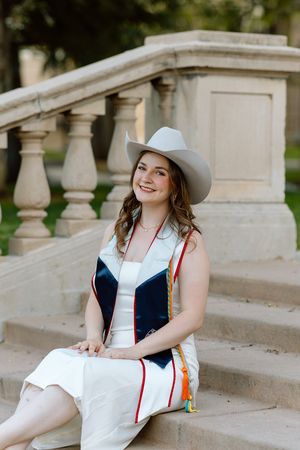 Smiling female graduate in white dress and cowboy hat wearing a navy graduation stole on stone steps.