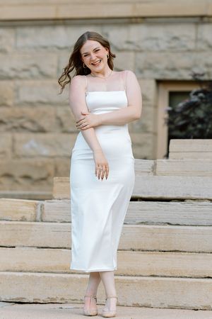Young woman in white midi dress poses on stone steps, smiling confidently at her graduation photoshoot.