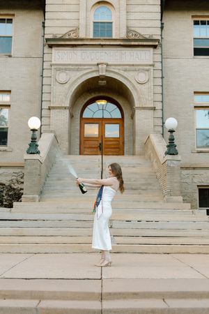 Graduate in white dress popping champagne bottle on steps of Simon Guggenheim Hall building.