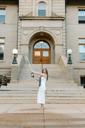 Joyful graduate in white dress celebrating with champagne bottle in front of Simon Guggenheim Hall steps.