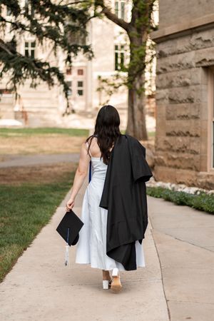 Graduate in white dress carrying black cap and gown walks along campus sidewalk past stone building.