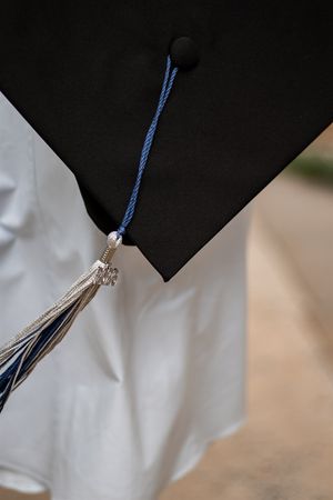 Close-up of a black graduation mortarboard cap with blue and silver tassel worn over a white graduation gown.