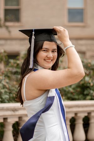 Happy female graduate in white dress adjusting black mortarboard cap with blue sash outdoors.