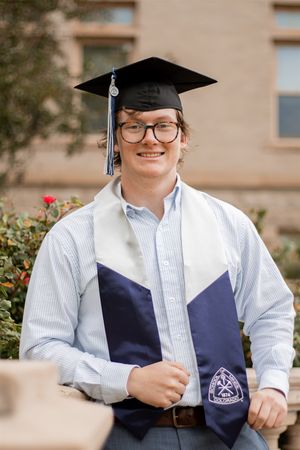 Smiling graduate wearing cap, stole, and glasses poses outdoors in front of brick building for graduation photo.