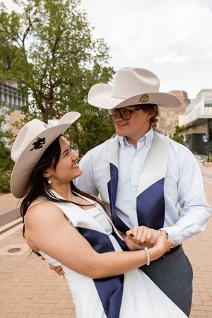 Couple in cowboy hats and graduation sashes smiling at each other outdoors on a brick plaza.