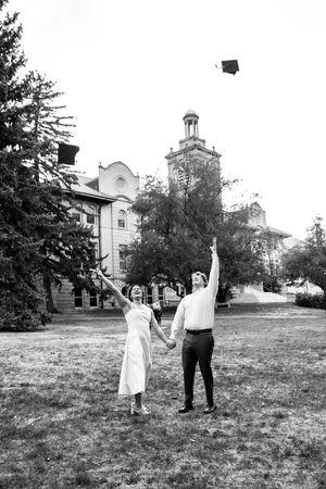 Black and white photo of a couple holding hands while tossing a graduation cap in front of a historic university building.