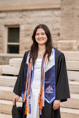 Smiling female graduate in black gown with blue and orange honor cords and stole posing outdoors.
