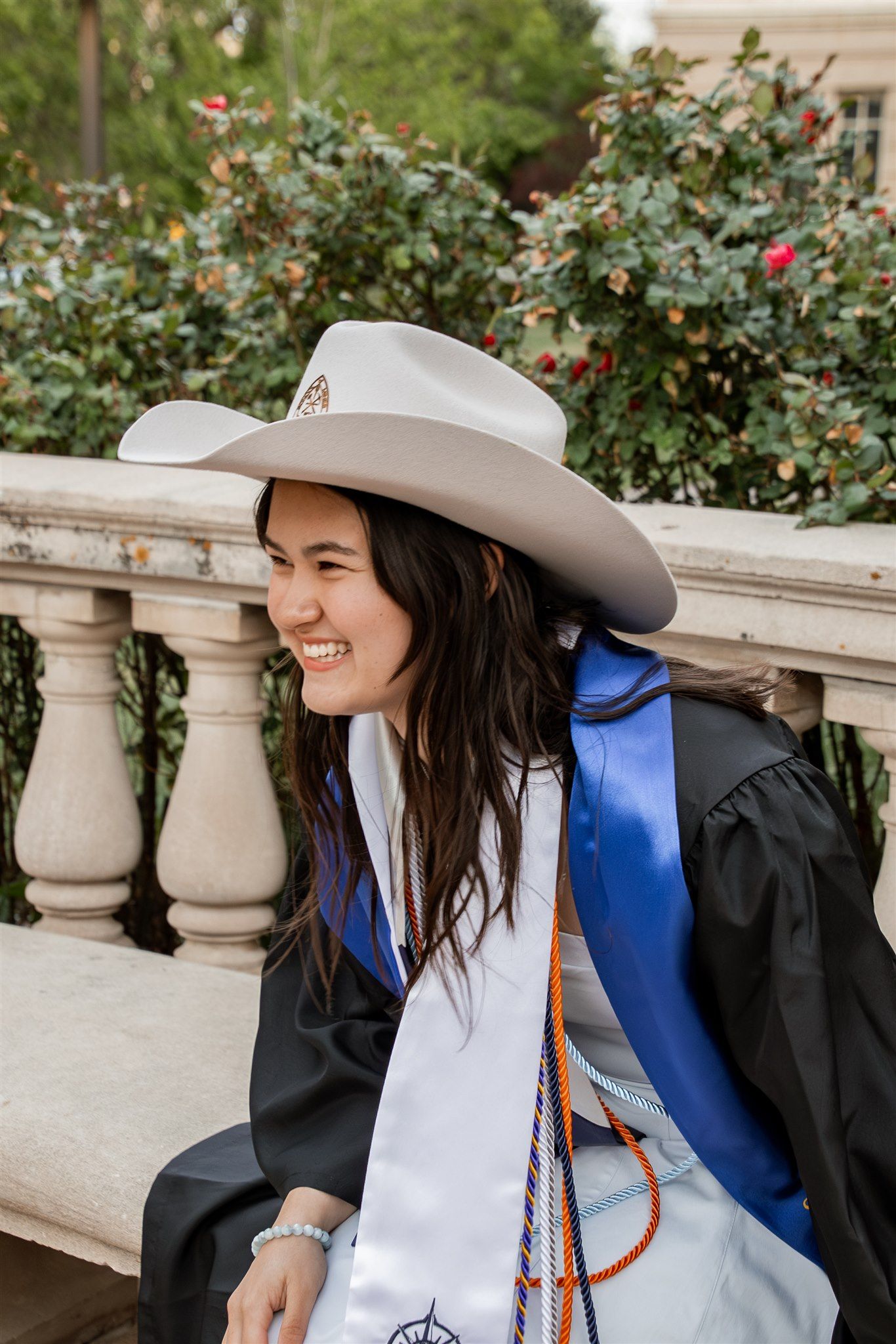 Smiling graduate in black gown and white cowboy hat sits by stone balustrade with roses.