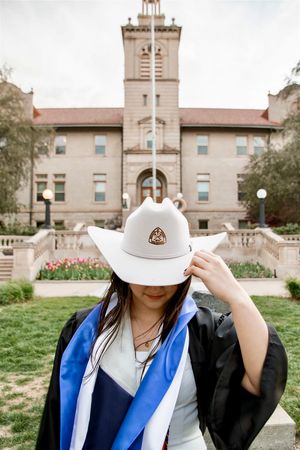 Graduate in black gown, white cowboy hat, and blue sash poses before historic university building.