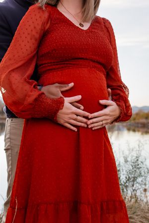 A woman in a red lace dress cradles her pregnant belly while standing in a natural outdoor setting.