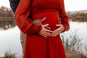Woman in red dress cradles her pregnant belly by the waterfront during sunset.