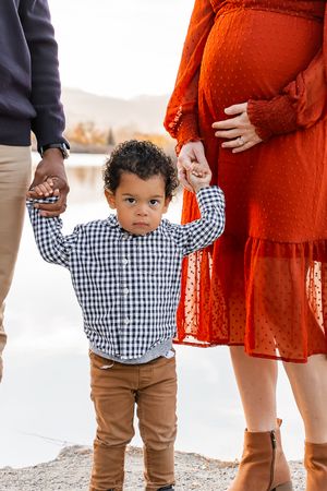 A small child in a gingham shirt and brown pants stands next to someone wearing a flowing rust-colored dress outdoors.