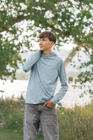 Teen boy in light blue hoodie and gray jeans standing outdoors near a tree, looking away thoughtfully.