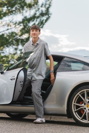 Young man in gray shirt and jeans leaning against a silver Porsche 911 sports car outdoors.