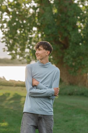 Young man in light blue turtleneck sweater standing outdoors with arms crossed, smiling, with green trees in background.