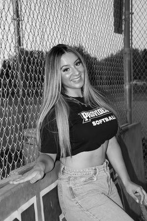 A black and white portrait against chain link fence wearing softball team apparel.