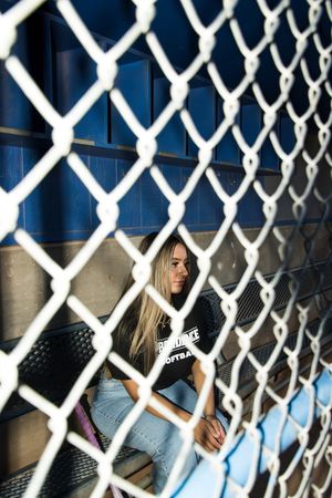 View through chain link fence of person in black t-shirt and jeans sitting on blue surface on sunny day.