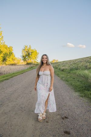 Woman in white floral maxi dress walking on dirt path with autumn trees in background.