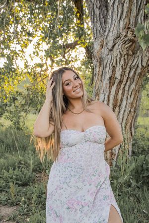 Woman in floral dress poses next to large tree trunk in natural outdoor setting.