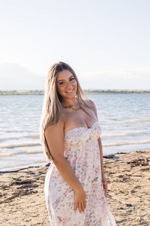 Woman in floral dress poses on beach at sunset with water and mountains in background.