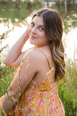 A young woman in a floral summer dress poses in a sunlit field of tall grass during golden hour.