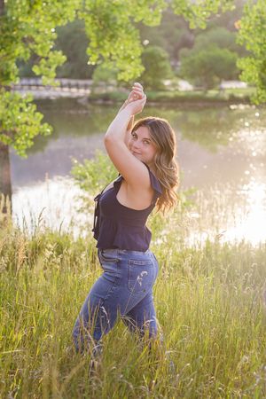 A figure poses in a field of tall grass at sunset near a tranquil lake, wearing jeans and a dark tank top.
