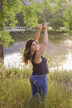 Person in dark tank top and jeans poses joyfully with raised arms in tall grass by a lake at sunset.