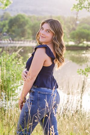 Young woman in navy top and jeans poses in tall grass by lake during golden hour sunset.