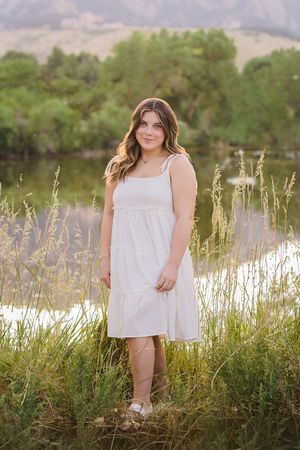 A white sundress-clad figure stands in tall grass by a lake during golden hour with mountains in the background.