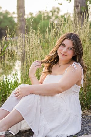 A person in a white summer dress sits among tall grasses at golden hour beside a lake.