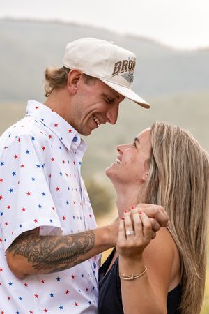 A smiling couple shares a joyful moment outdoors, the man wearing a star-print shirt and cap, the woman with long blonde hair.