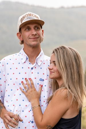 Couple posing outdoors, man in star-print shirt and Broncos cap, woman showing engagement ring on his chest.