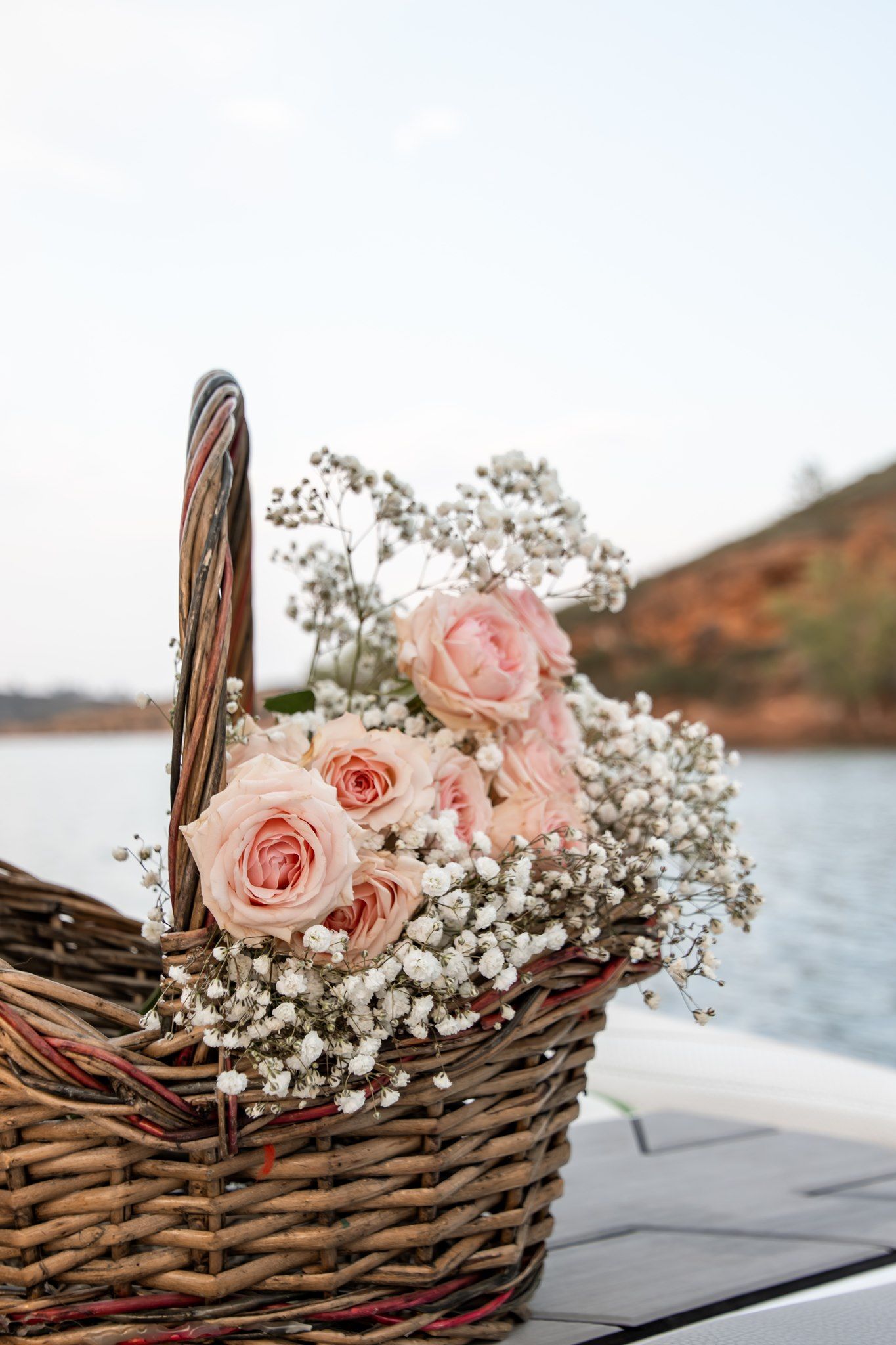 A wicker basket filled with pink roses and white baby's breath flowers on a boat with a lake backdrop.