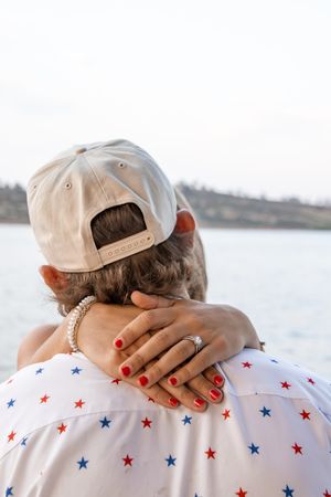 Back view of couple embracing near water, woman with red nails and engagement ring resting hand on mans shoulder.
