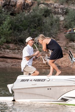 Man proposes on a boat bow to surprised woman near rocky cliffs on a lake, SR450 visible on hull.