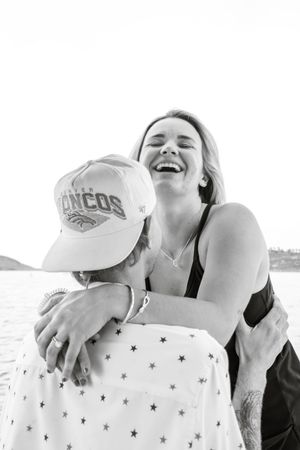 A joyful woman laughing while embracing a man wearing a Denver Broncos cap near a waterfront.