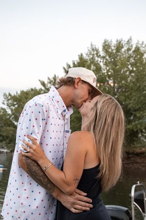 A couple shares a romantic kiss near a lake, the man wearing a star-print shirt and white cap.
