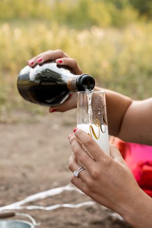 A woman with red nails pours champagne into a flute, wearing an engagement ring outdoors.
