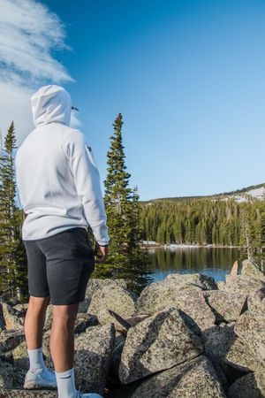 Person in white hoodie overlooking mountain lake surrounded by evergreen forest.
