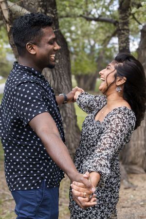 A joyful couple shares a laugh while holding hands among trees near a lake.